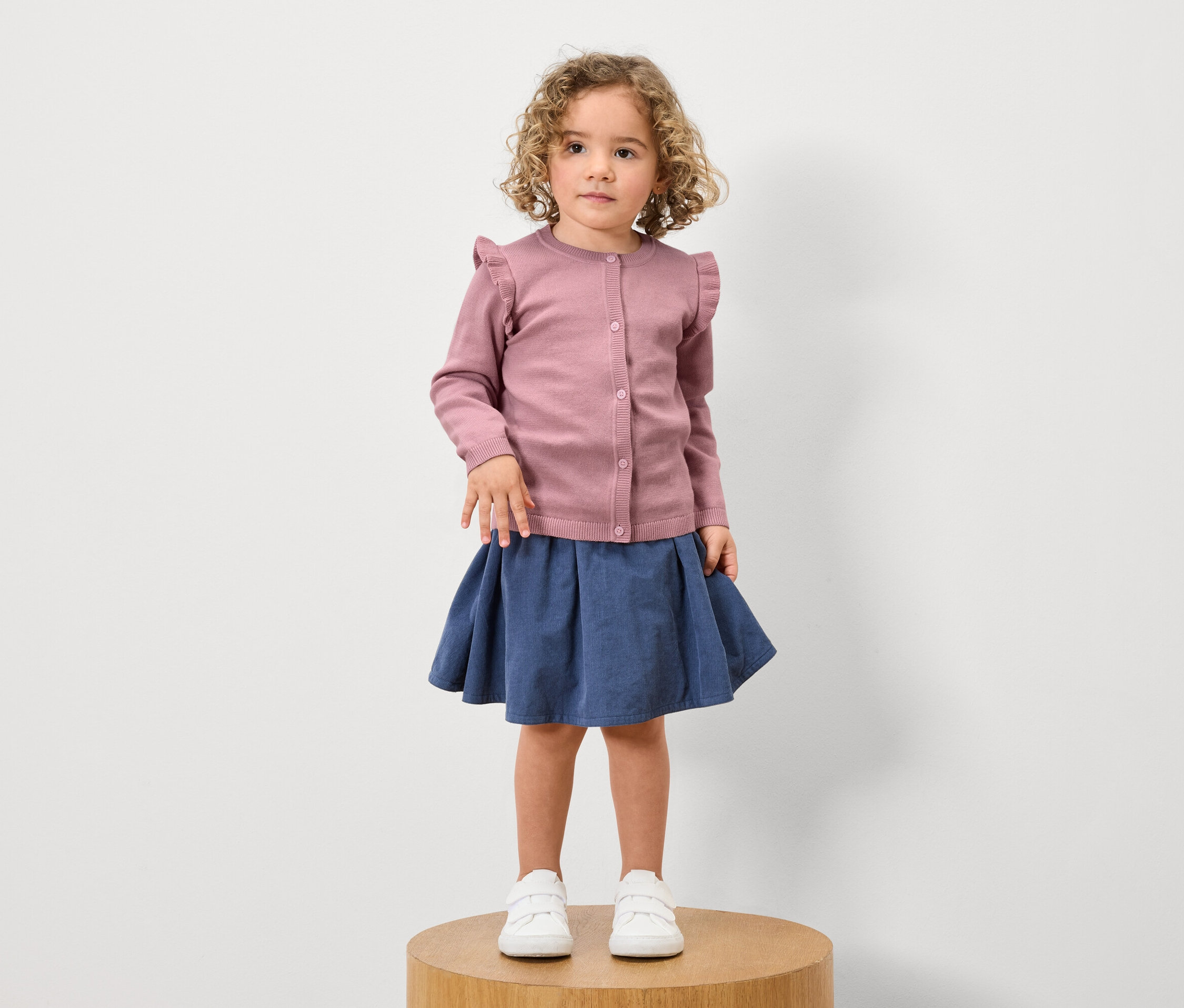 Une fille aux cheveux bouclés se tient sur un socle en bois et porte un cardigan à volants rose, une jupe bleue et des baskets blanches.