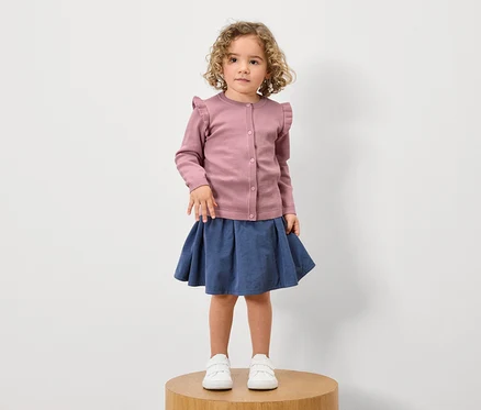 Une fille aux cheveux bouclés se tient sur un socle en bois et porte un cardigan à volants rose, une jupe bleue et des baskets blanches.