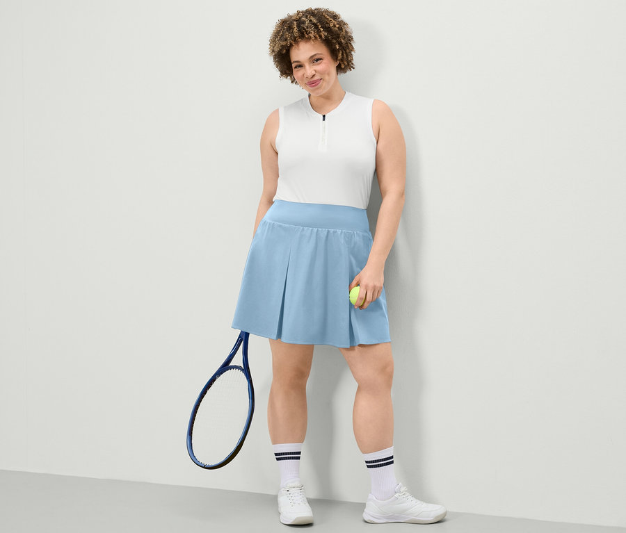 Femme posant dans un top fonctionnel blanc et une jupe de sport bleu clair. Elle tient une balle de tennis et une raquette de tennis.