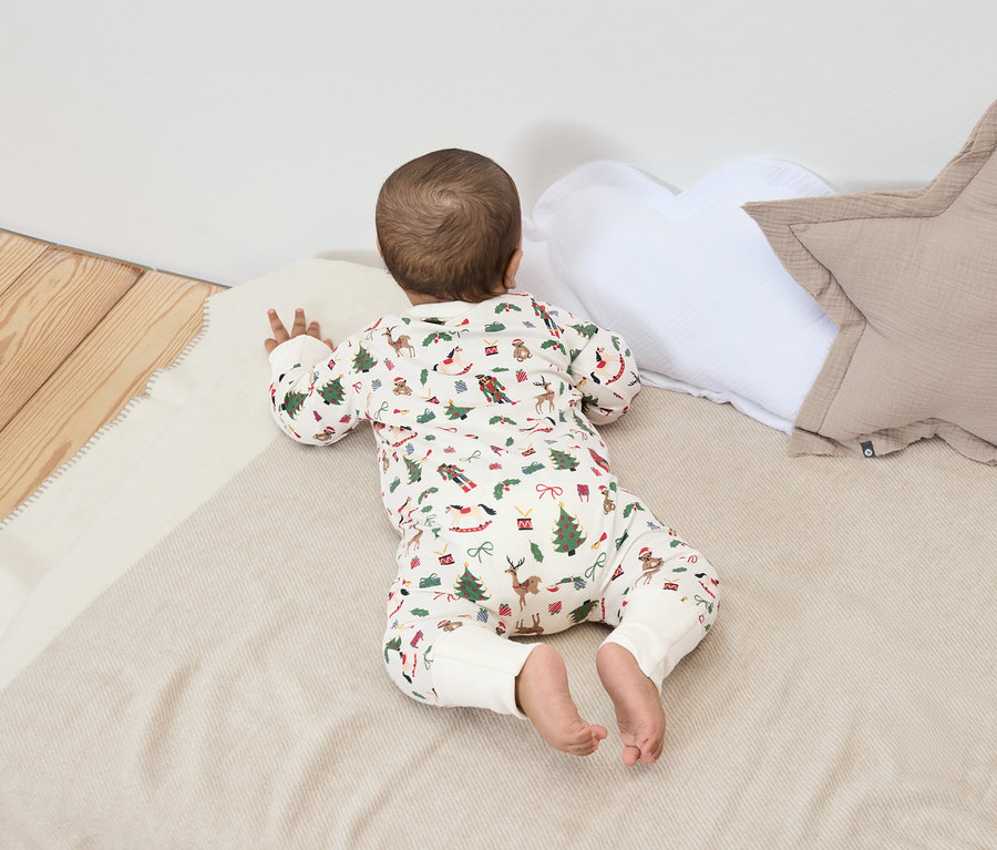 Un bébé est allongé sur le ventre sur une couverture à motifs de Noël, à côté d'un oreiller blanc et beige.