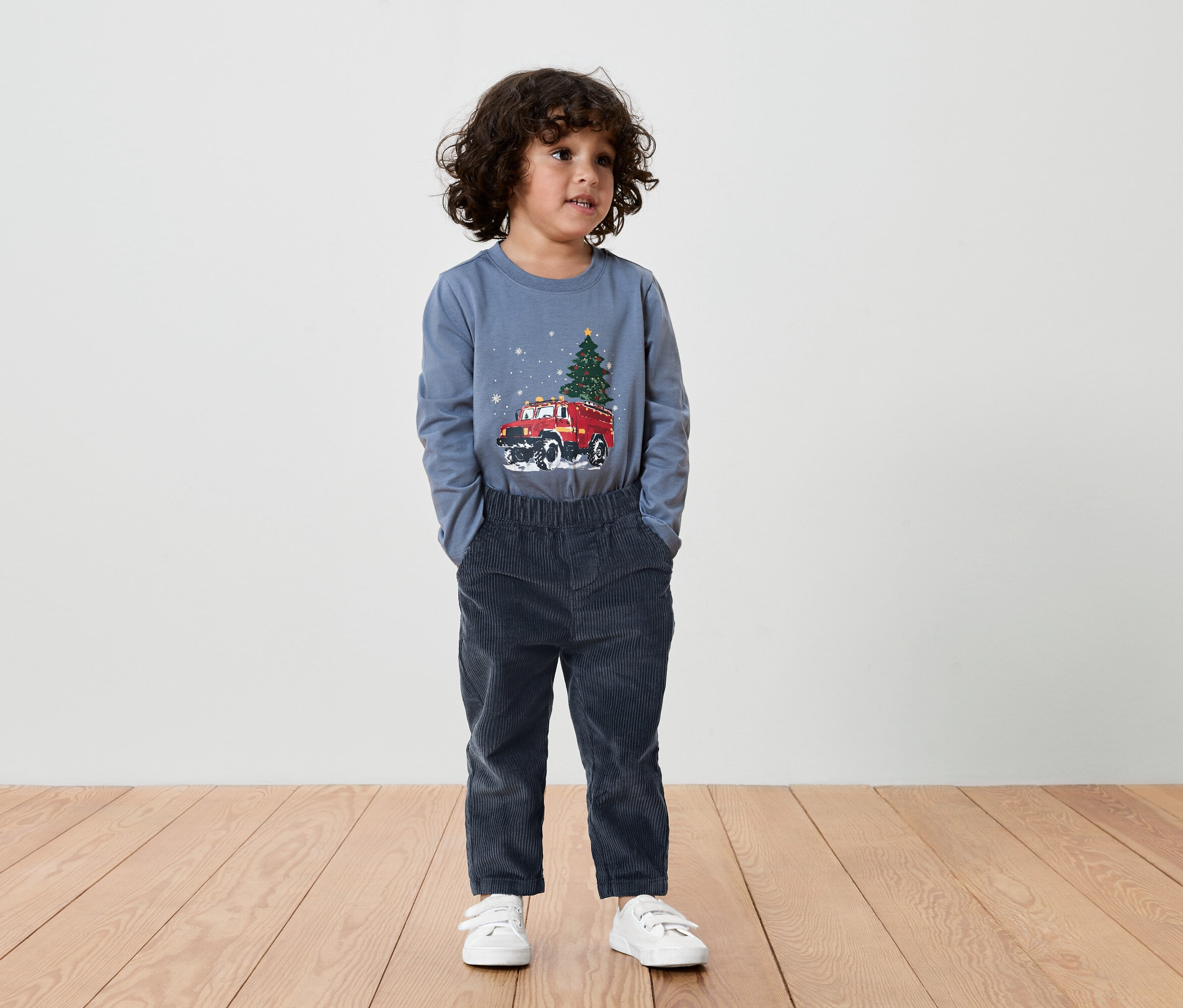 Un garçon aux cheveux bruns porte un T-shirt bleu avec une voiture rouge et un sapin de Noël imprimés dessus, ainsi qu'un pantalon bleu foncé et des baskets blanches.