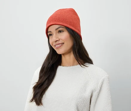 Une femme aux longs cheveux bruns et souriante porte un bonnet orange et un pull blanc.
