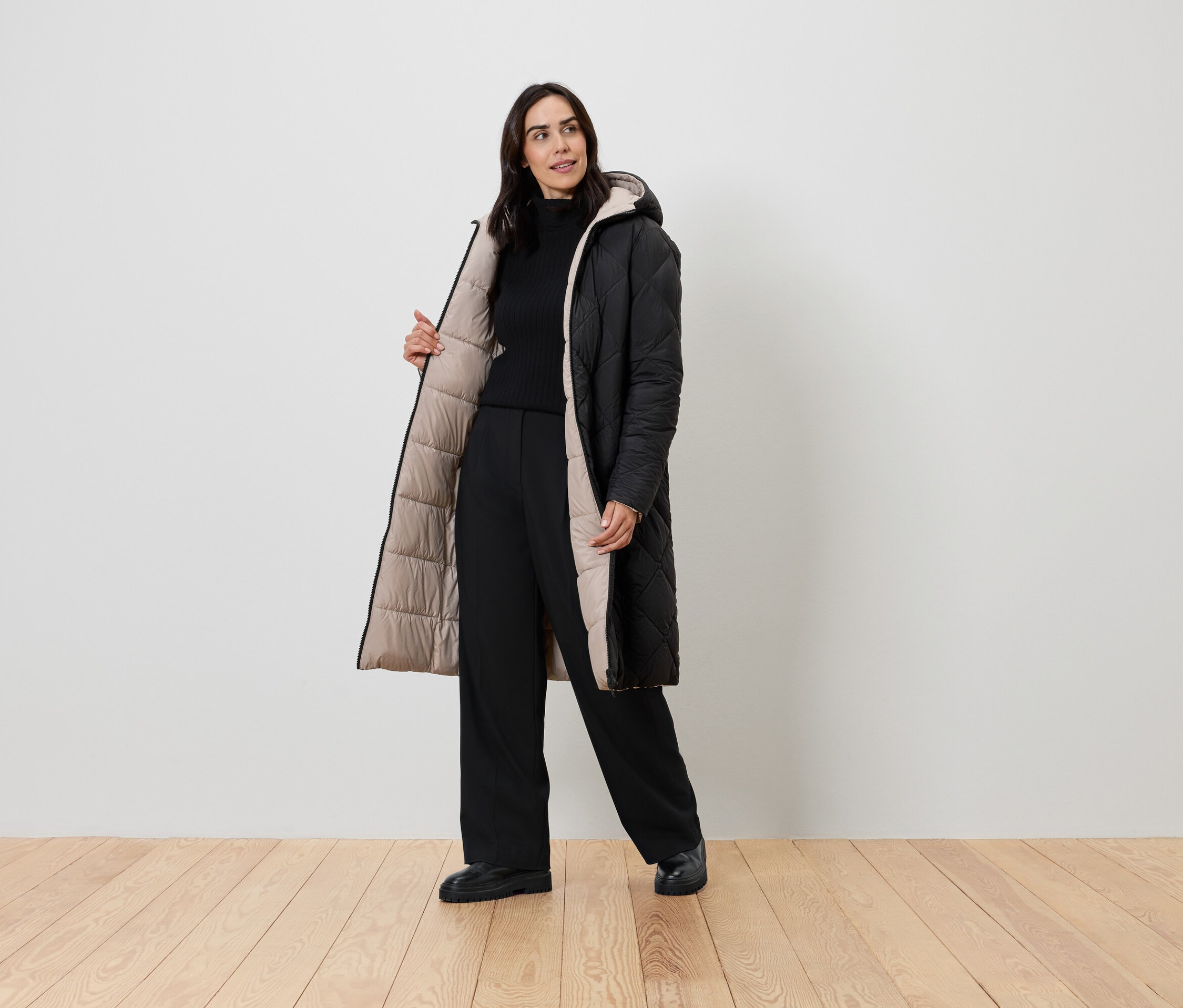 Une femme pose dans un manteau noir avec une doublure beige, un pull à col roulé noir et un pantalon noir.