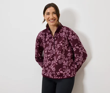 Une femme souriante porte un sweat-shirt en polaire bordeaux à fleurs et un pantalon foncé.