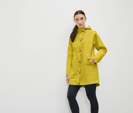 Une femme portant un manteau de pluie jaune à capuche et un legging noir se tient devant un mur blanc.