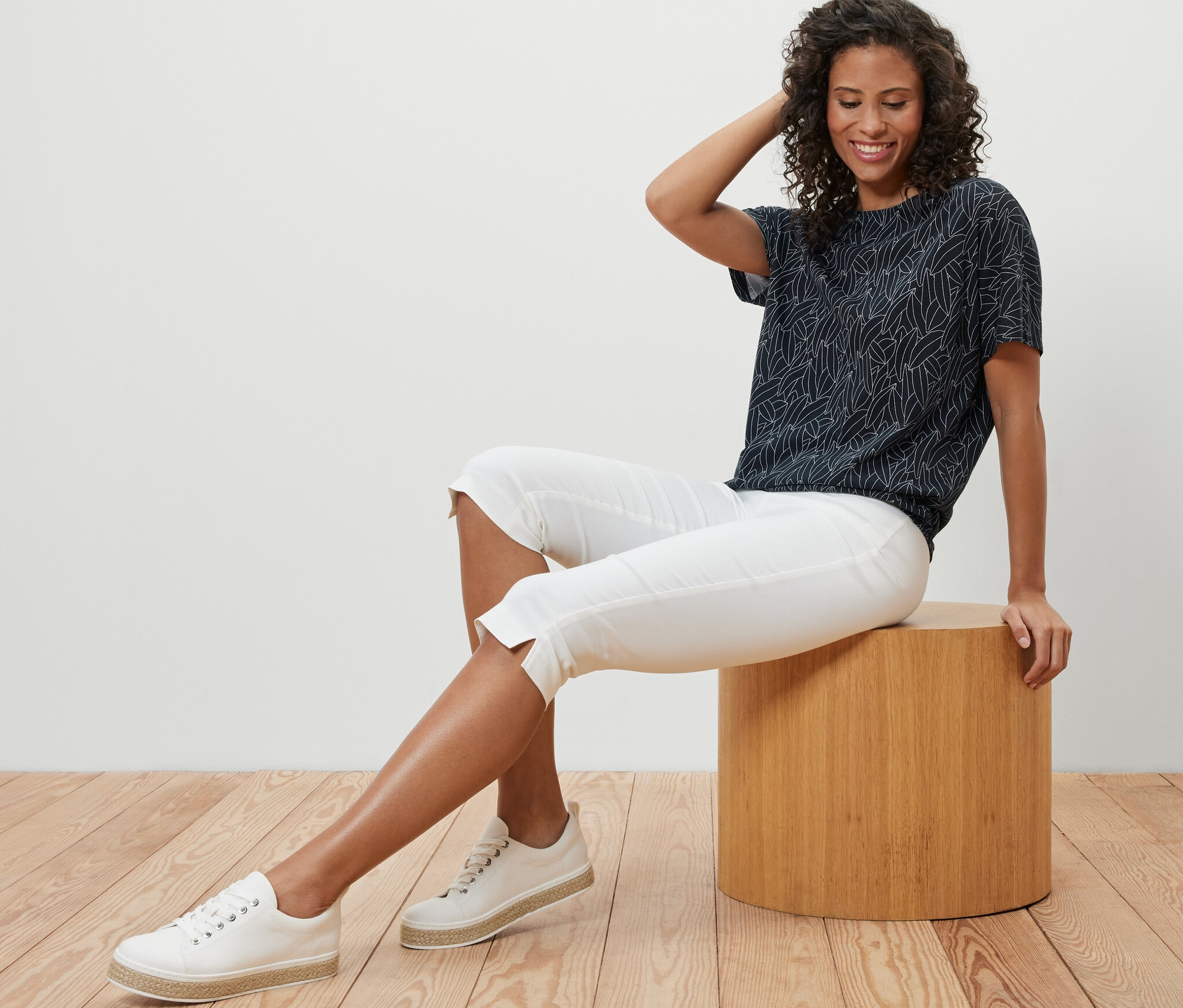 Une femme est assise sur un tabouret et porte un chemisier bleu foncé à encolure dégagée, un pantacourt blanc et des baskets.