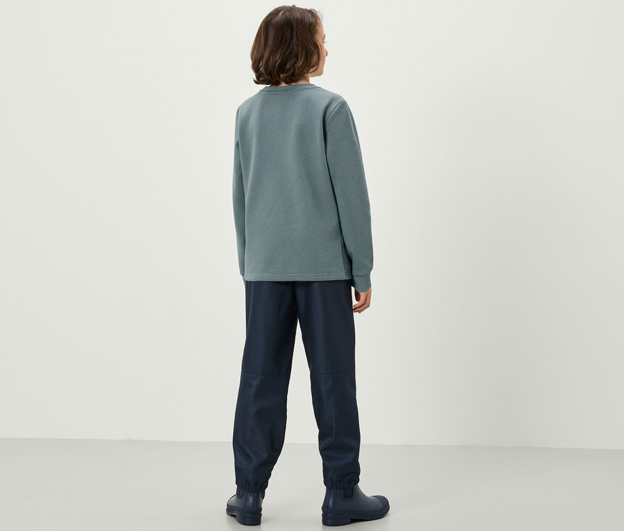 Un garçon se tient dos à la caméra portant un sweat-shirt bleu et sauge et un pantalon bleu foncé.