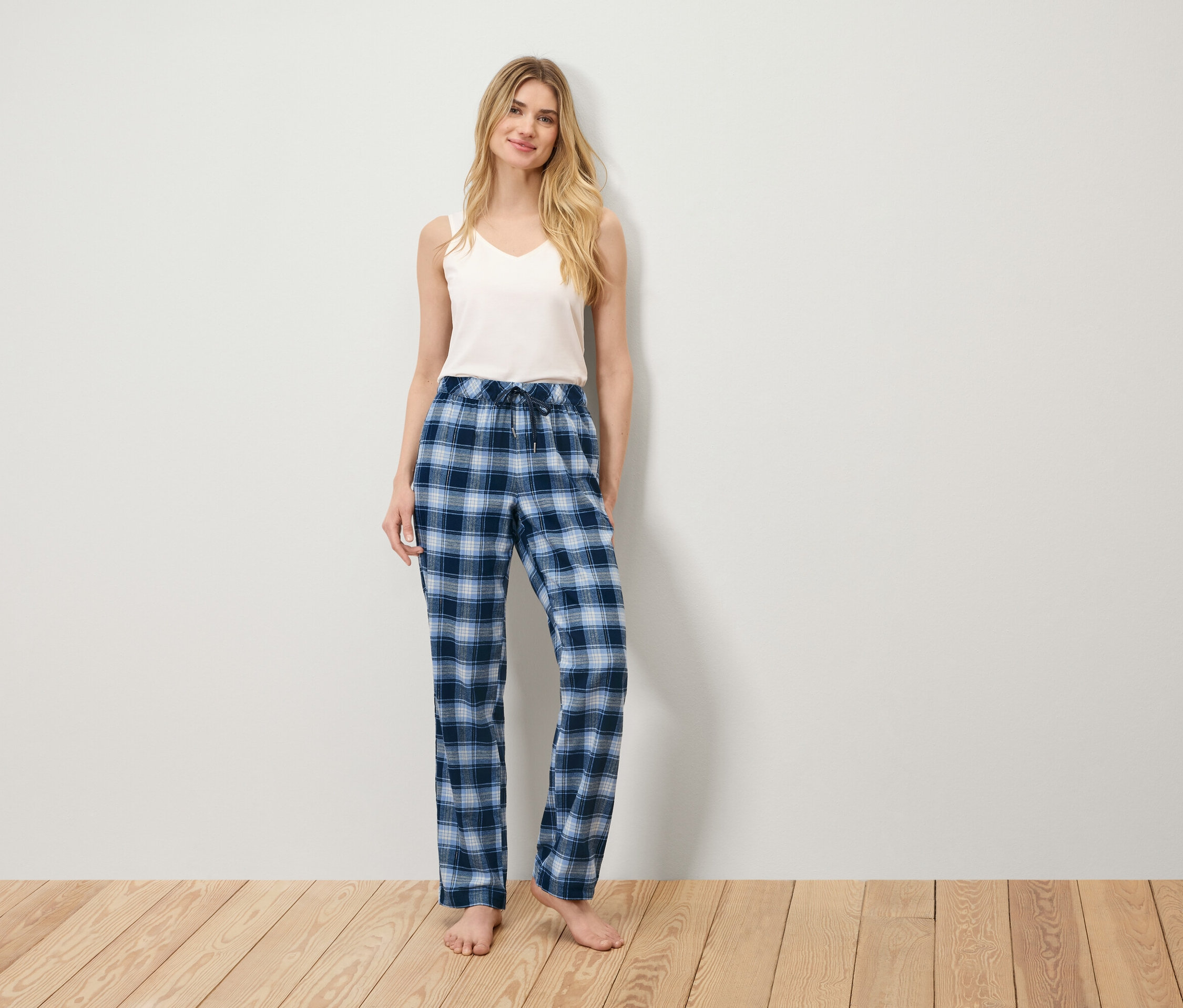 Une femme se tient sur un plancher en bois, portant un débardeur blanc et un pantalon de pyjama à carreaux bleus.
