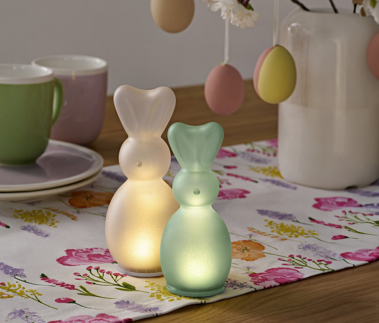 Deux figurines de lapins lumineux sont posées sur une nappe à motifs floraux, à côté de tasses, d'assiettes et d'un bouquet d'œufs de Pâques dans un vase.