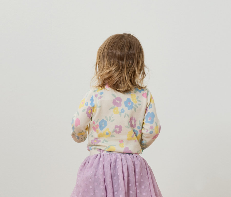 Petite fille vue de dos, portant un cardigan à fleurs et une jupe violette.