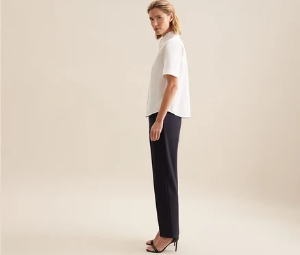 Femme debout de profil, regardant par-dessus son épaule, portant un chemisier à manches courtes Seidensticker blanc, un pantalon foncé et des sandales noires à talons.