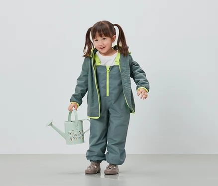 Fillette en pantalon et veste de pluie gris tenant un arrosoir vert clair.