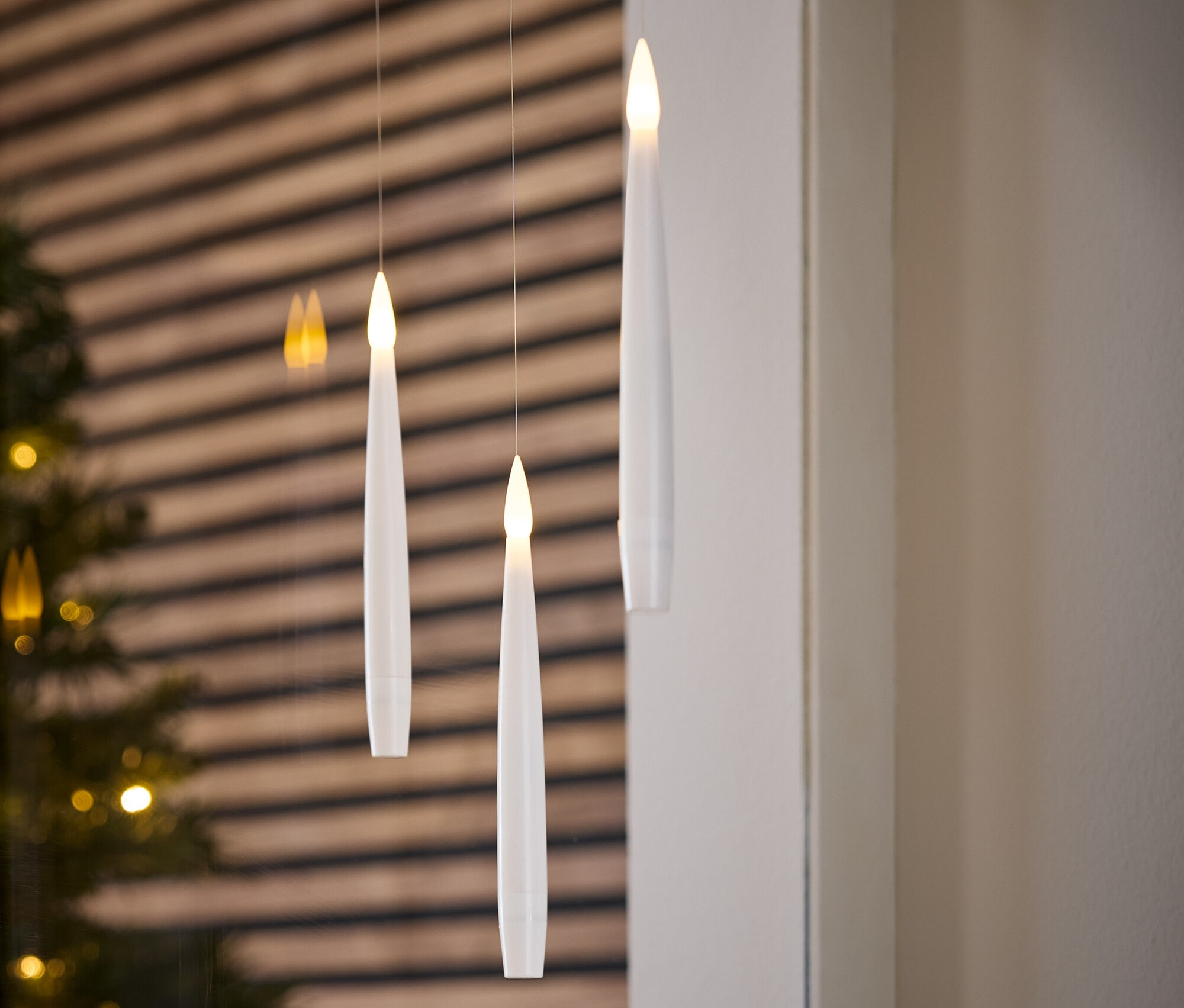 Trois bougies blanches électroniques suspendues à des fils. Un arbre de Noël et un mur en bois sont visibles en arrière-plan.