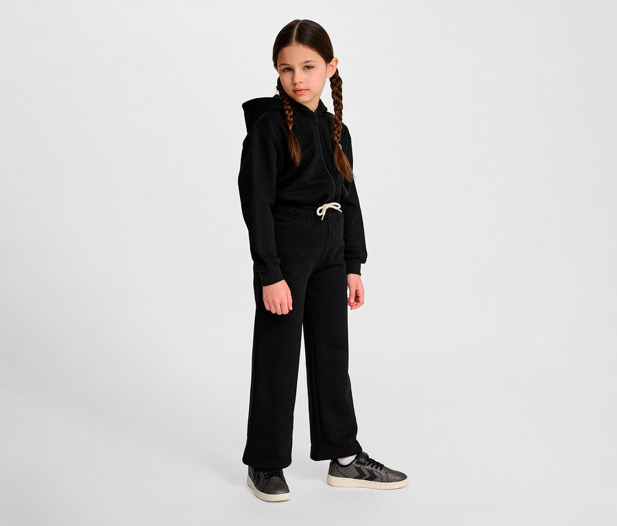 Une jeune fille avec des tresses se tient devant un fond et porte un sweat à capuche noir et un pantalon de survêtement ample HUMMEL HMLJR Loose WL.
