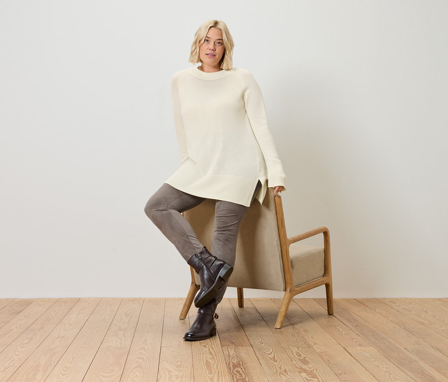 Une femme blonde pose, portant un long pull en tricot crème et s'appuyant sur une chaise.