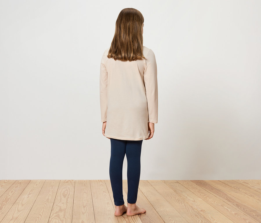 Une fille se tient dos à la caméra, portant une tunique crème et des leggings bleu marine.