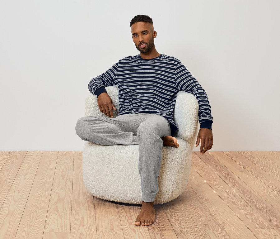 Un homme est assis dans un fauteuil. Il porte un t-shirt rayé à manches longues et un pantalon de survêtement gris.