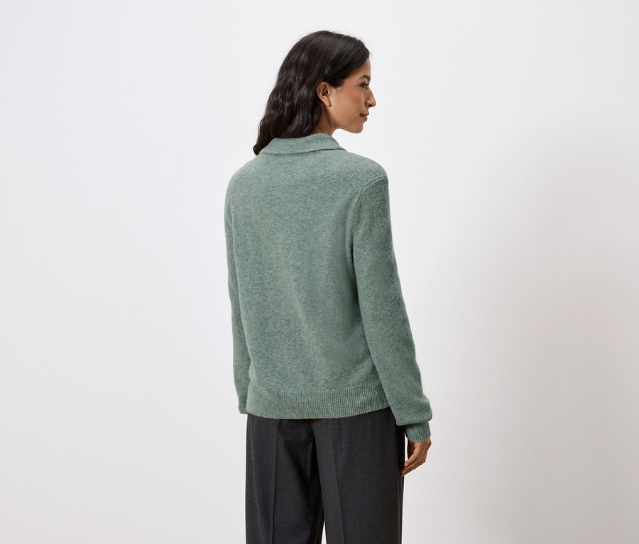 Vue de dos d'une femme aux cheveux foncés portant un pull vert et un pantalon foncé.