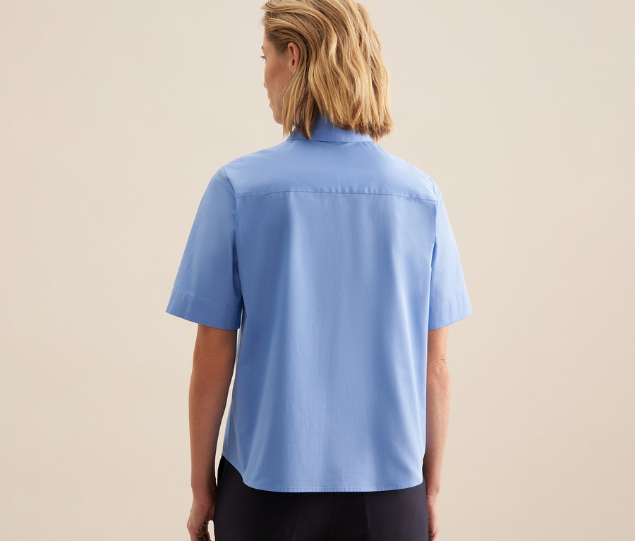 Femme vue de dos, portant un chemisier à manches courtes Seidensticker bleu clair et un pantalon foncé.