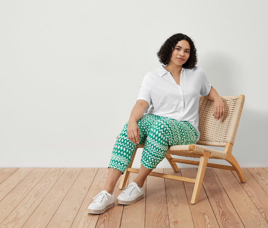 Une femme est assise sur une chaise et porte un pantalon 3/4 en jersey imprimé et une blouse blanche avec patte de boutonnage.