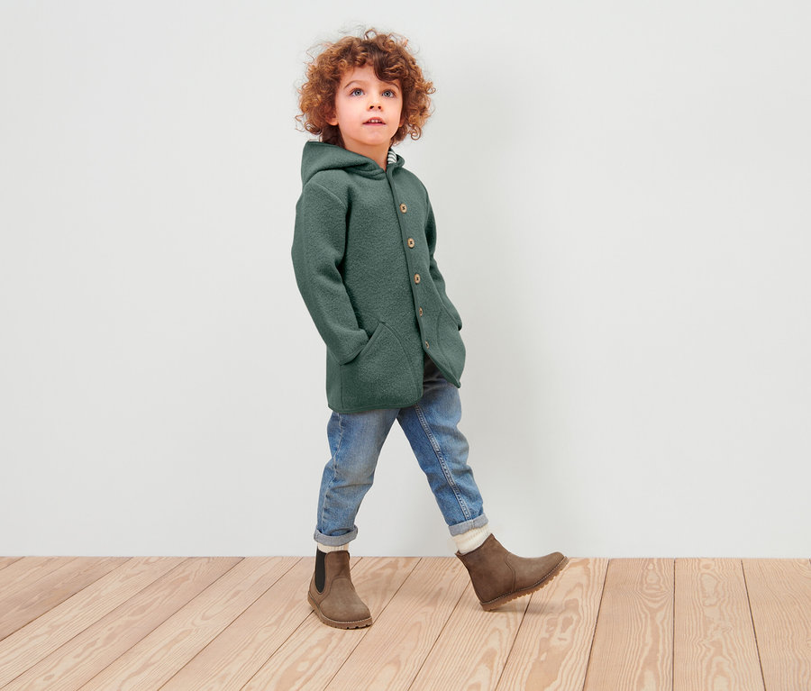 Un garçon aux cheveux bouclés marche sur un plancher en bois, portant un manteau en laine vert à capuche, un jean et des bottes marron.