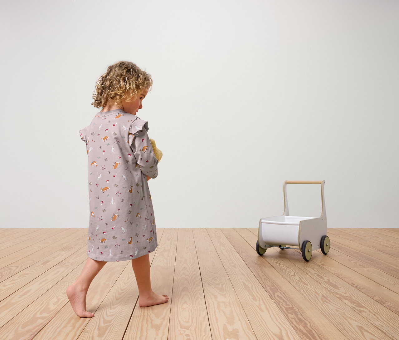 Fille vue de dos portant 2 chemises de nuit pour enfants tenant une peluche. Un chariot à roulettes jouet est posé sur un plancher en bois.