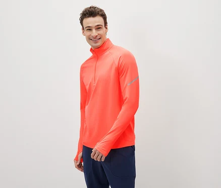 Un homme souriant pose, vêtu d'un haut de sport corail à manches longues et d'un pantalon de survêtement bleu marine.