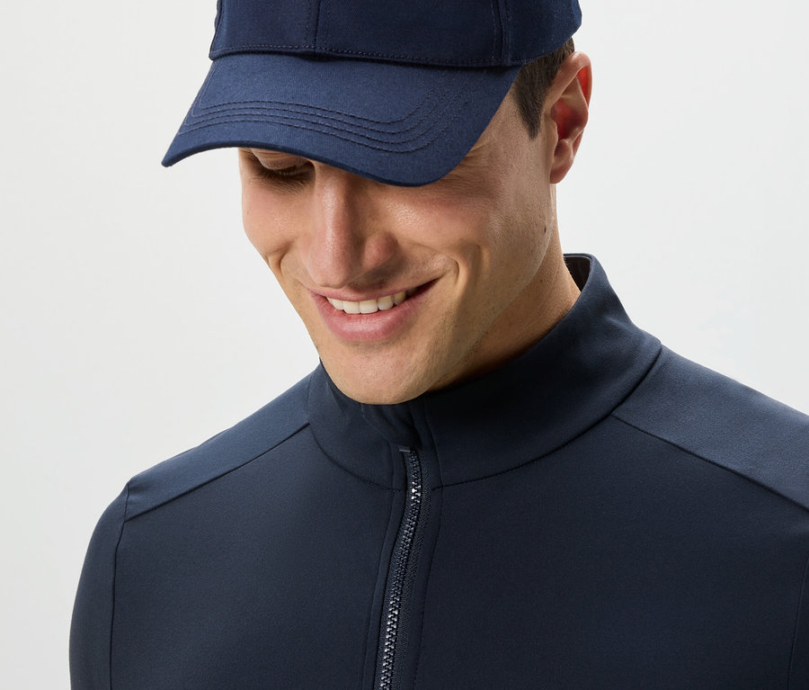 Homme en veste fonctionnelle bleu marine et casquette regardant vers le bas.