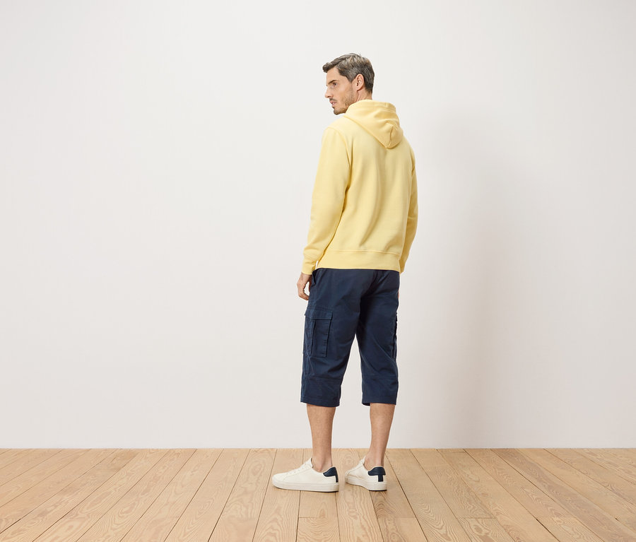 Homme vu de dos, portant un sweat à capuche jaune, un short bleu et des baskets blanches.