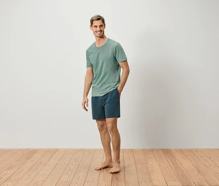 Un homme se tient pieds nus sur un plancher en bois, portant un t-shirt vert et un short bleu.