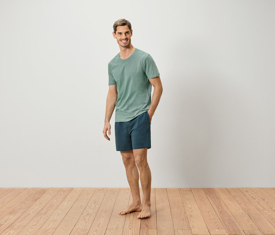 Un homme se tient pieds nus sur un plancher en bois, portant un t-shirt vert et un short bleu.