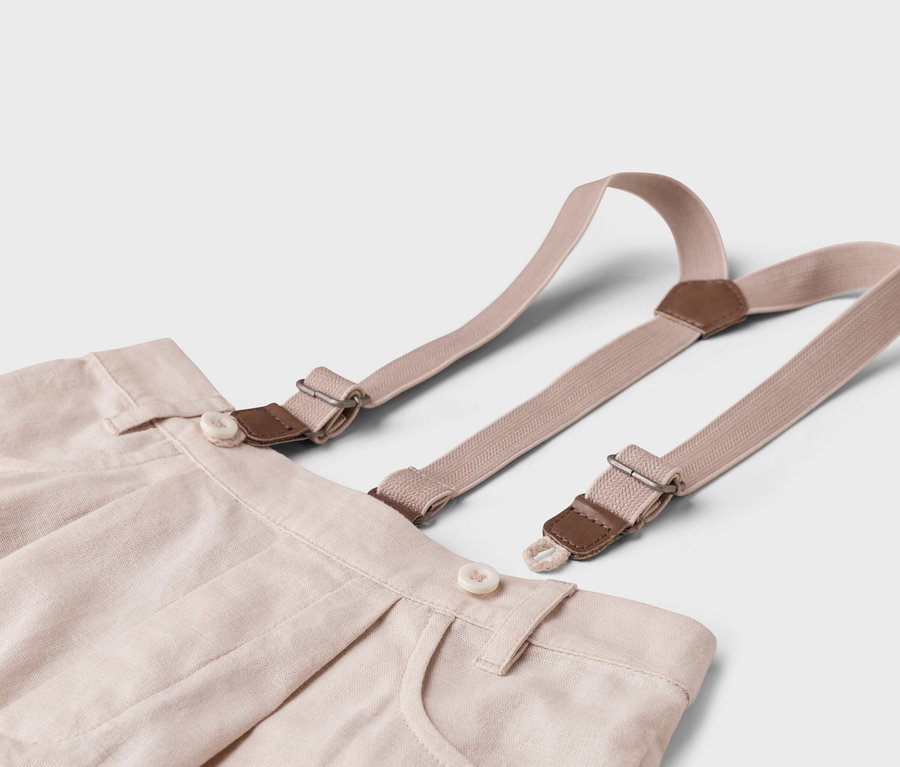 Gros plan d'un pantalon en tissu beige avec des bretelles sur fond blanc.