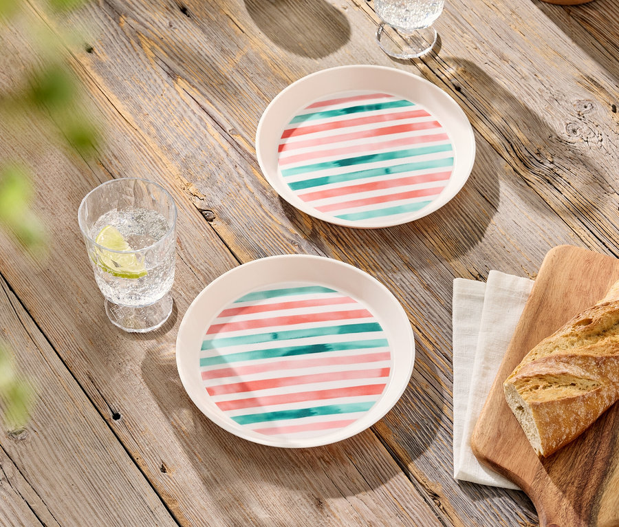 Deux assiettes à rayures roses et turquoise, deux verres avec des boissons, des olives dans un bol et du pain sur une planche à découper sur une table en bois.