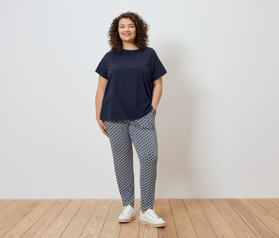 Une femme se tient devant un mur, portant un t-shirt bleu marine, un pantalon à motifs et des chaussures blanches.