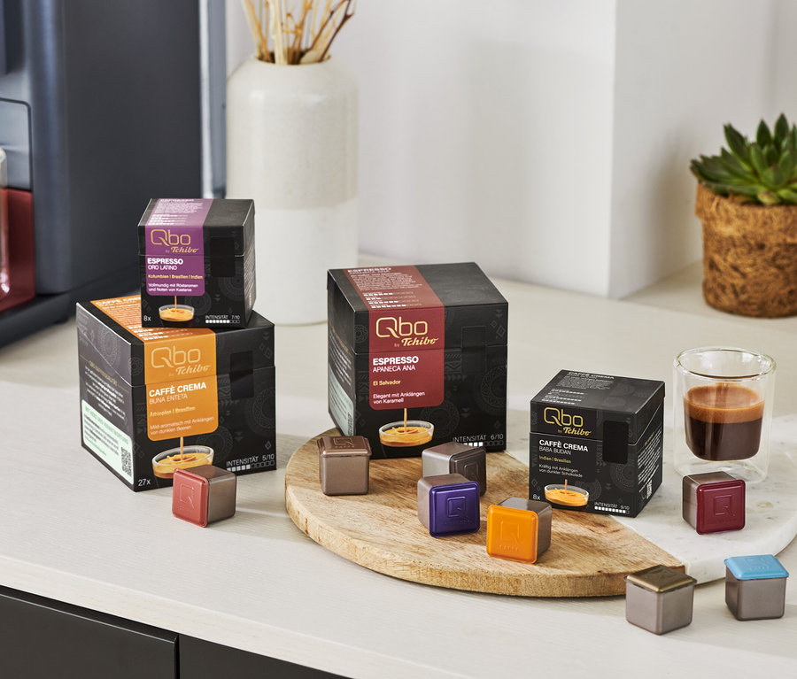 Sur une table, il y a des capsules Qbo Espresso APANECA ANA et une machine à café avec un expresso dans une tasse.