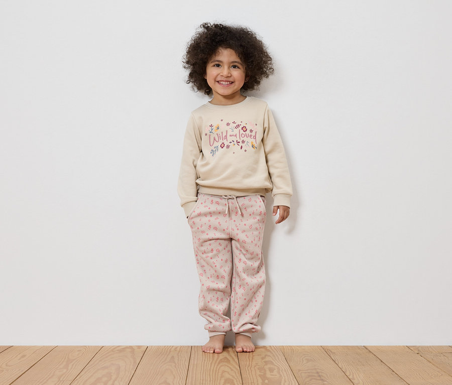 Une fillette souriante aux cheveux bouclés se tient pieds nus devant un mur blanc, vêtue d'un pull beige avec l'inscription "Wild and loved" et d'un pantalon rose.
