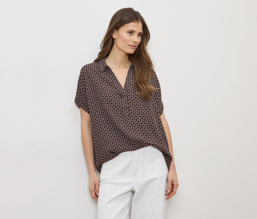 Femme portant une chemise marron à motifs et un pantalon blanc sur fond blanc.