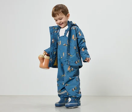 Un garçon souriant vêtu d'un manteau de pluie bleu et d'une salopette à motifs animaliers tient un arrosoir orange.