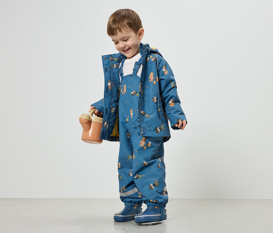 Un garçon souriant vêtu d'un manteau de pluie bleu et d'une salopette à motifs animaliers tient un arrosoir orange.