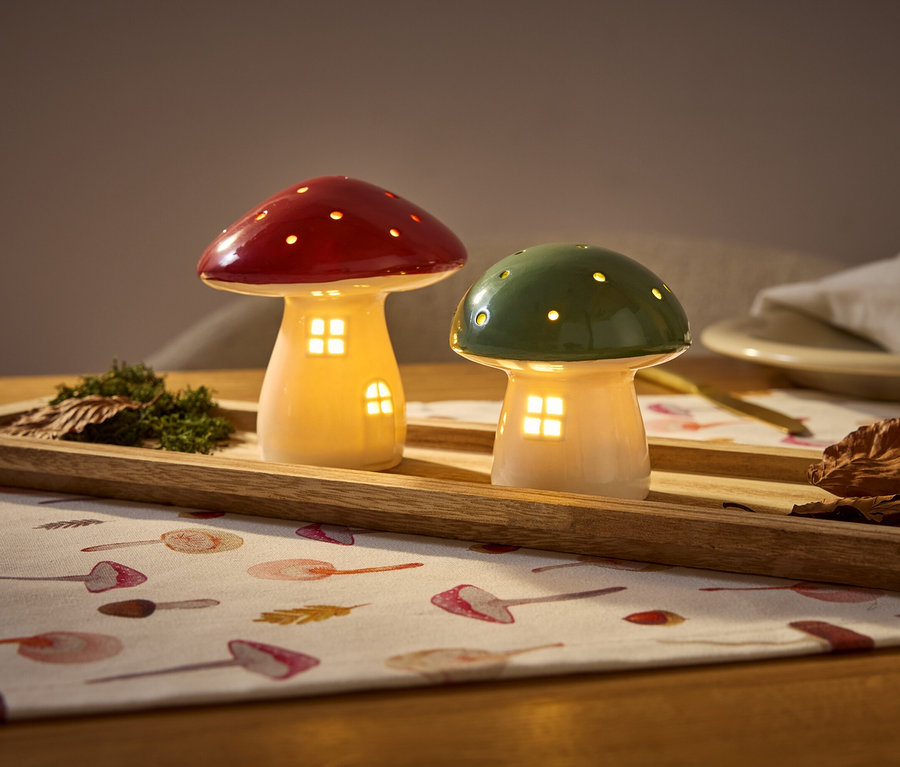 Deux maisons champignons lumineuses, une rouge et une verte, sont posées sur un plateau en bois avec de la mousse et des feuilles sur une nappe.
