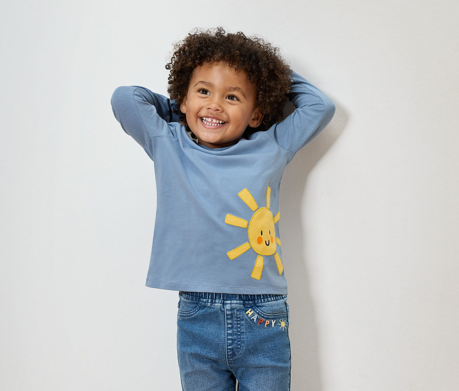 Un garçon souriant aux cheveux bouclés porte un t-shirt bleu à manches longues avec un soleil jaune et un jean bleu.