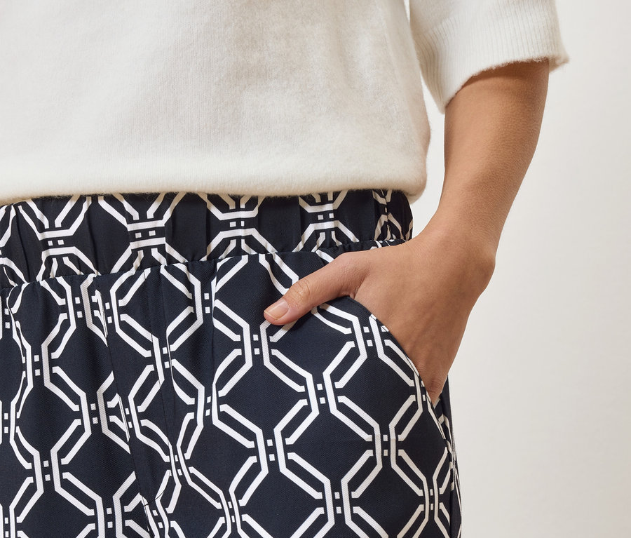 Gros plan d'une personne portant un pantalon bleu marine à motif géométrique blanc et un pull blanc, avec la main dans la poche.