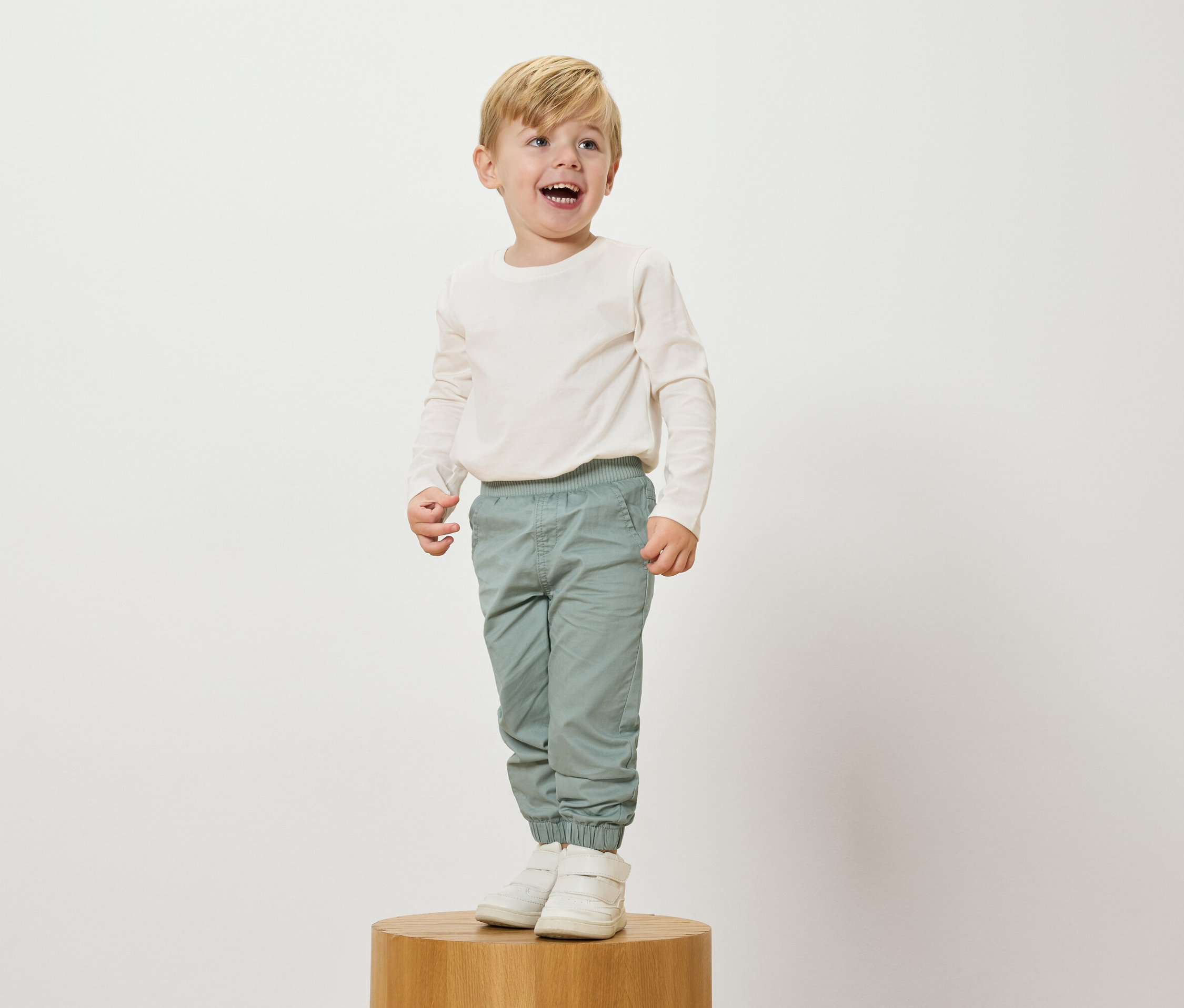 Un garçon blond souriant se tient sur un socle en bois, portant un haut crème à manches longues, un pantalon bleu clair et des baskets blanches.