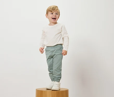 Un garçon blond souriant se tient sur un socle en bois, portant un haut crème à manches longues, un pantalon bleu clair et des baskets blanches.
