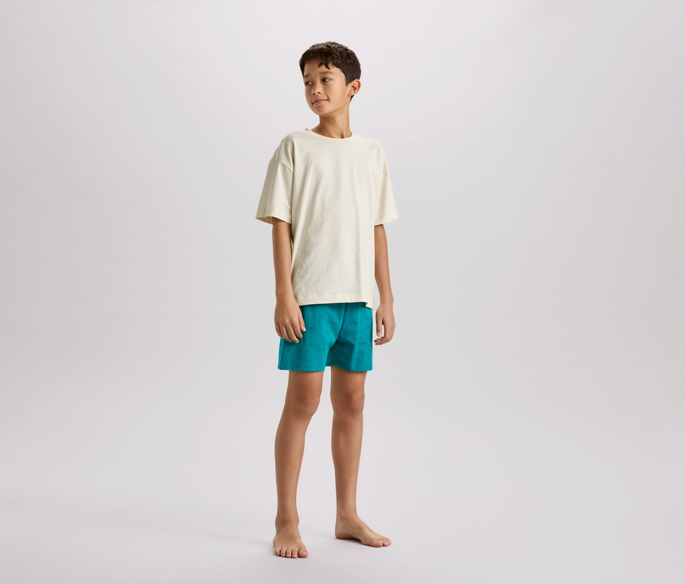 Un garçon pieds nus se tient de côté, portant un t-shirt crème et un short turquoise.