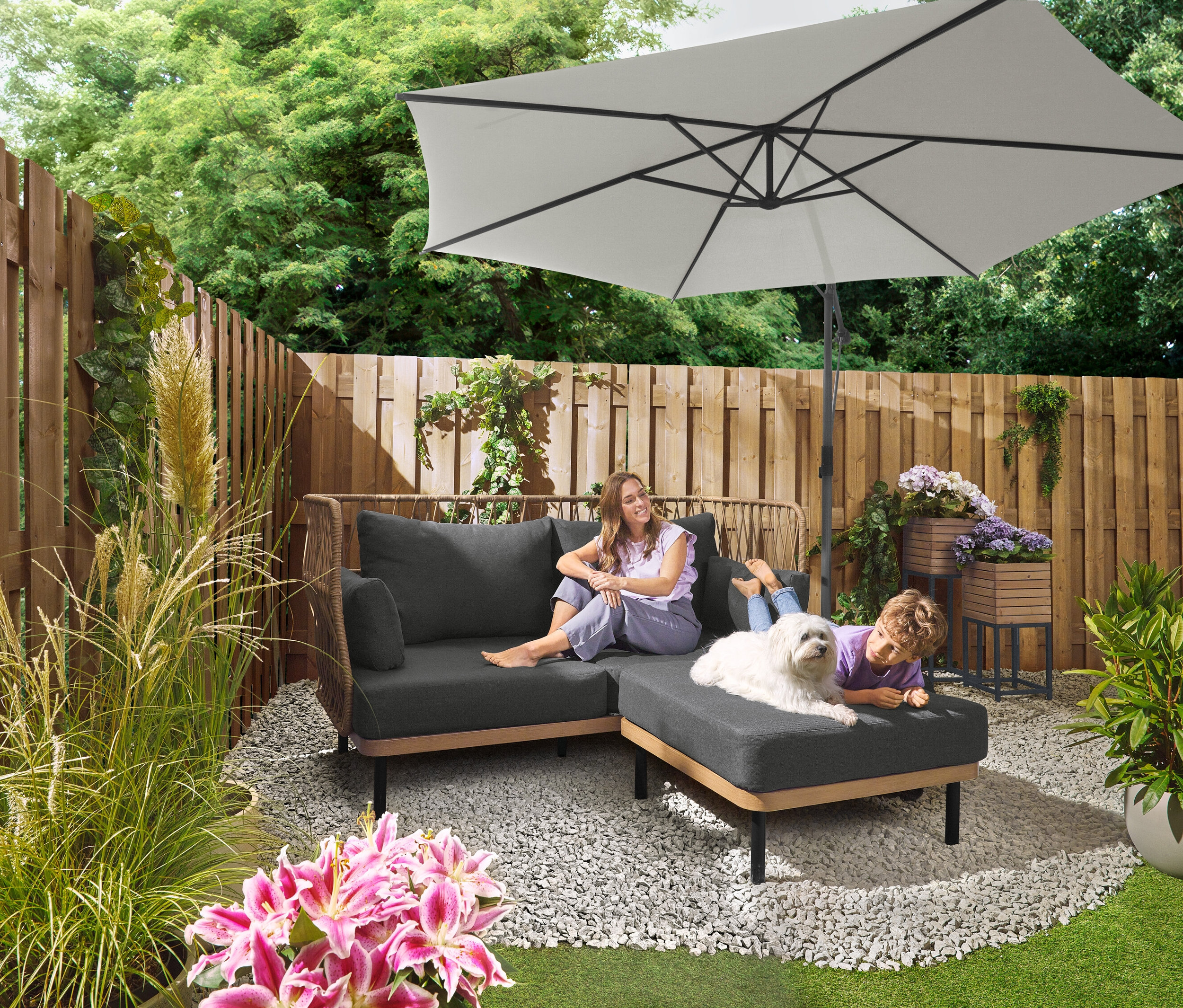 Une femme est assise sur un canapé gris sous un parasol Leco Ampelschirm Ø 300 cm avec support et housse de protection. Un enfant est allongé avec un chien sur un ottoman. À l'arrière-plan, on peut voir un ensemble de plaques Leco pour pied en croix.