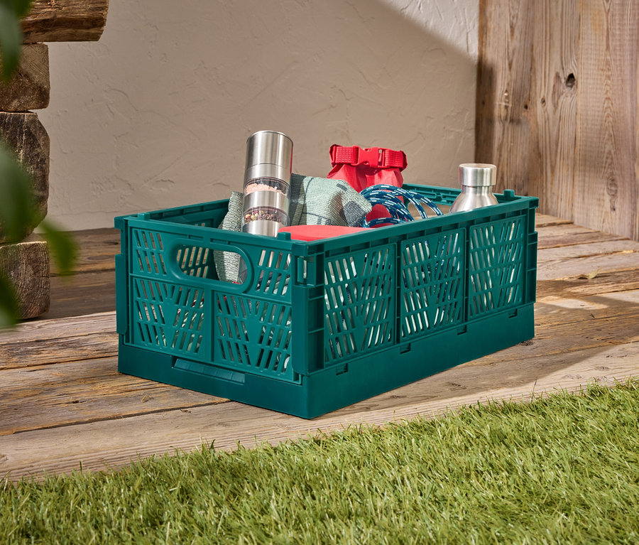 Une boîte en plastique verte est posée sur un plancher en bois, remplie d'un moulin à poivre en acier inoxydable, d'un sac rouge, d'une corde bleue et d'une bouteille en acier inoxydable.