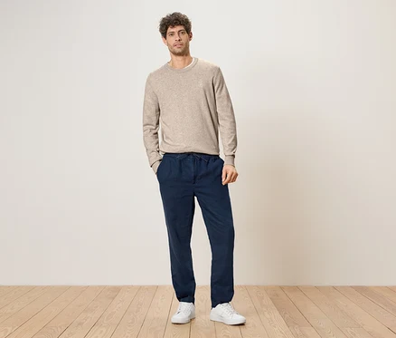 Un homme aux cheveux bouclés se tient les mains dans les poches devant un mur, portant un pull marron clair, un pantalon bleu foncé avec un cordon de serrage et des baskets blanches.