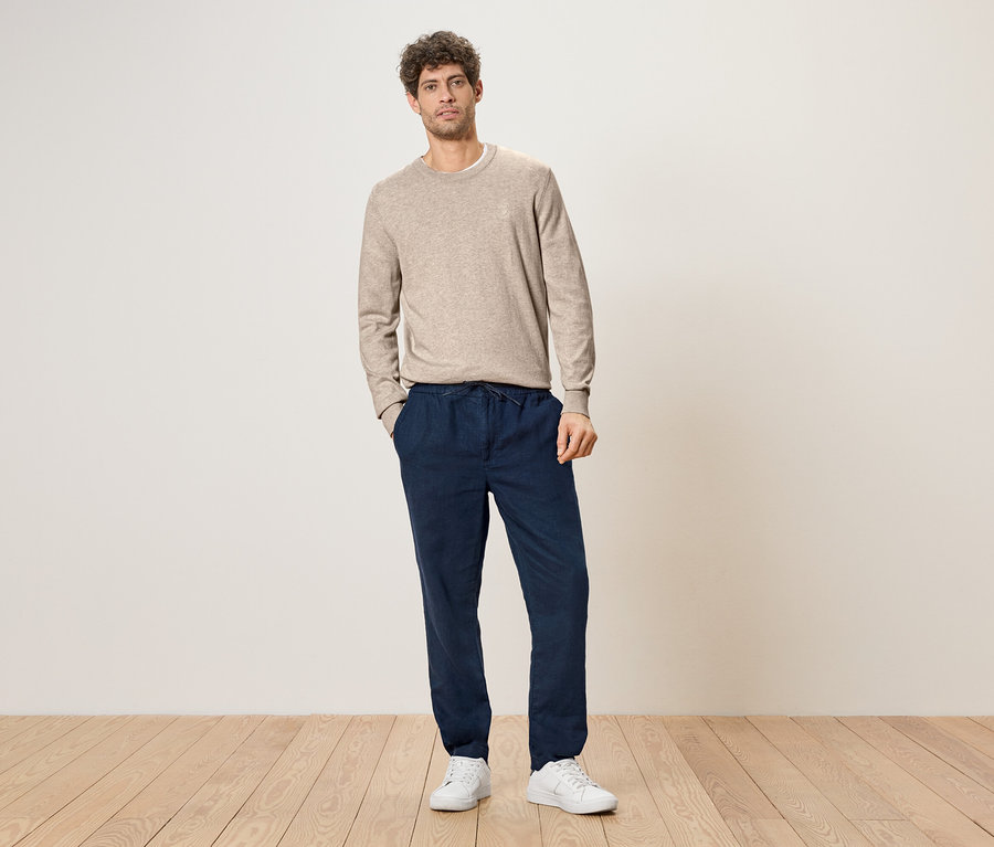 Un homme aux cheveux bouclés se tient les mains dans les poches devant un mur, portant un pull marron clair, un pantalon bleu foncé avec un cordon de serrage et des baskets blanches.