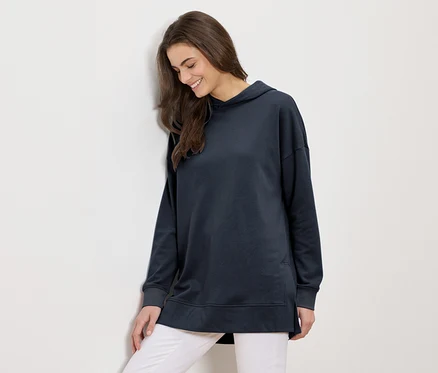 Une femme aux longs cheveux bruns portant un long sweat à capuche bleu marine et un pantalon blanc sourit et s'appuie contre un mur blanc.